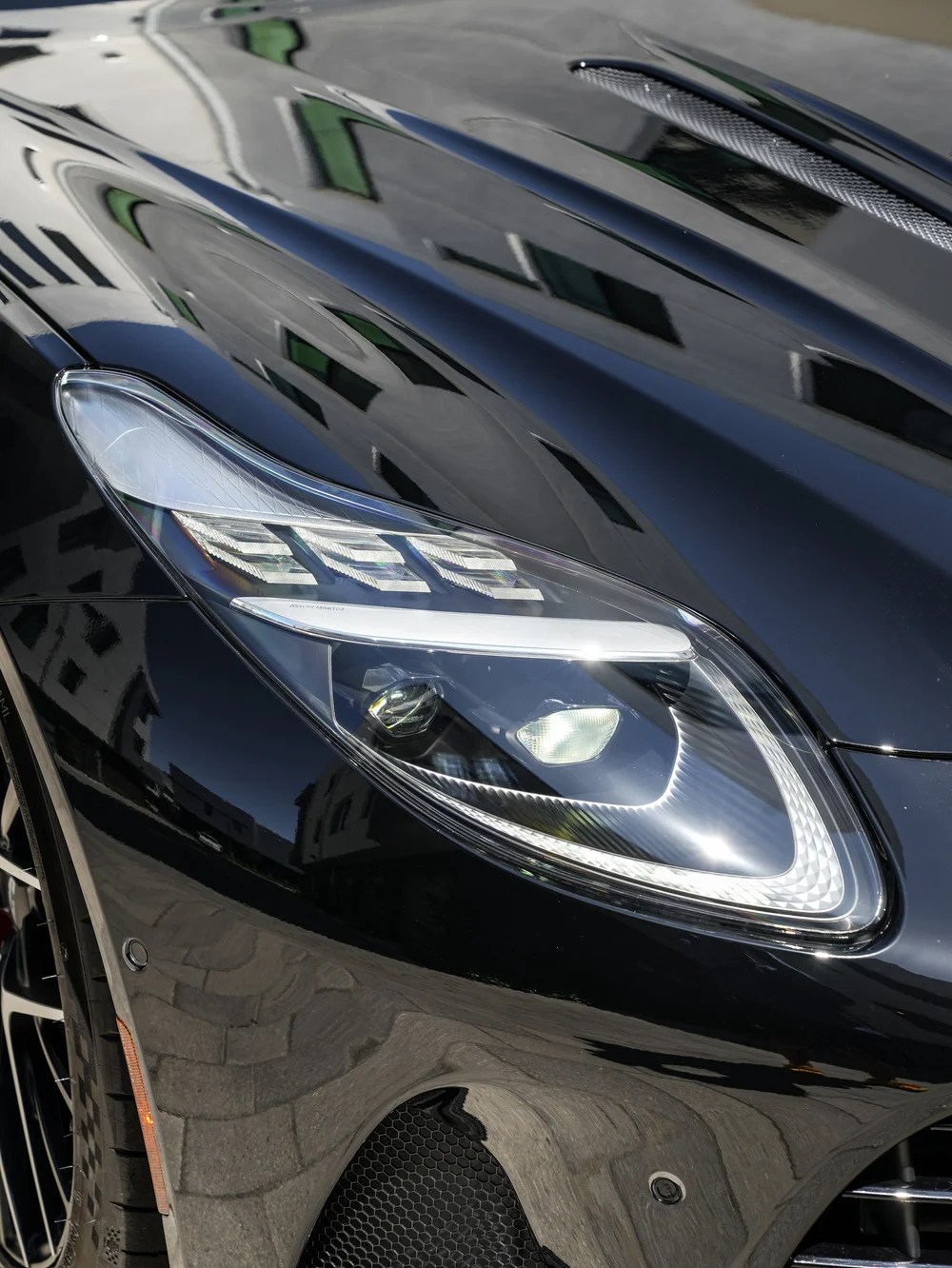 Illuminated DB12 headlamp beside reflective black hood paint and a hood vent.