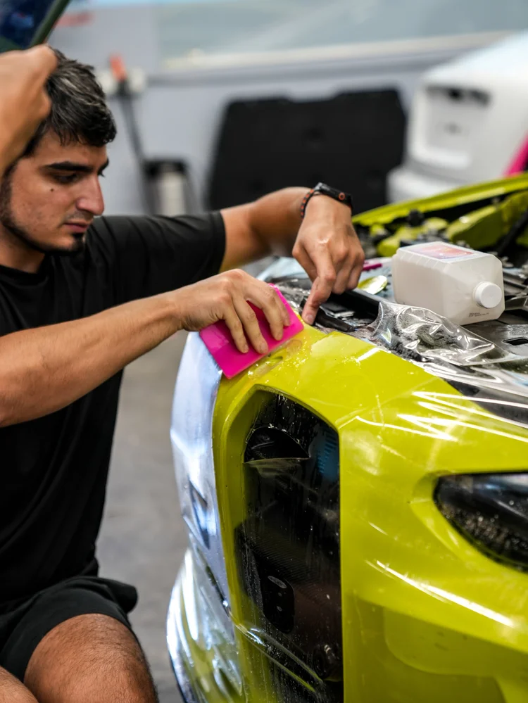 Technician using a pink flat tool against transparent film at an upper BMW M4 front edge beside a headlight and grille, with the hood raised in the background.