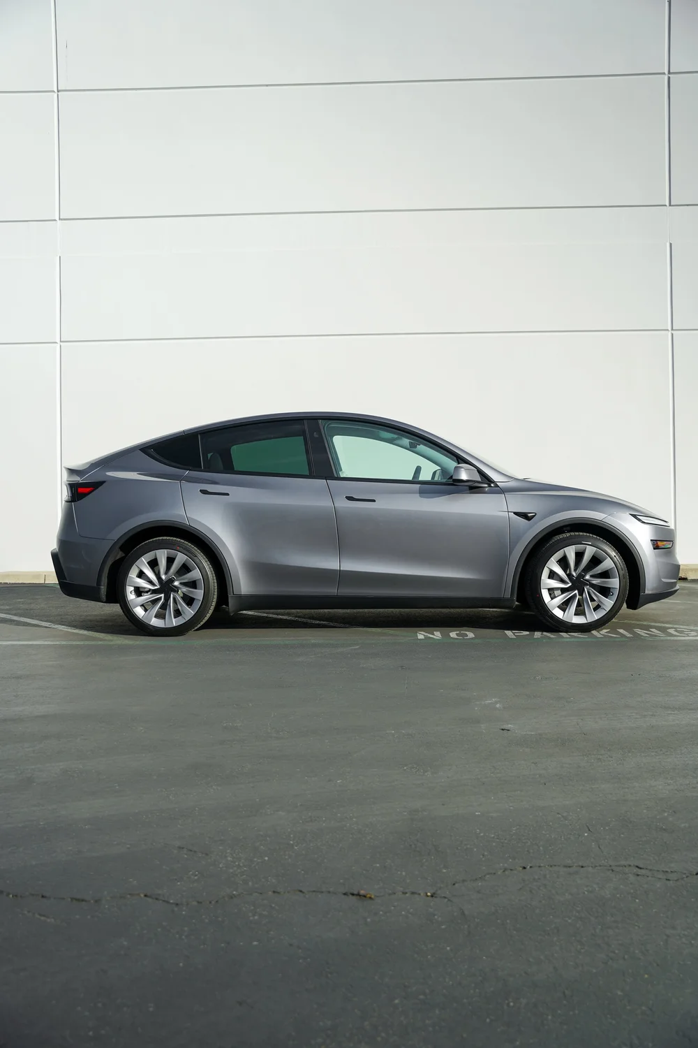 Complete side profile of the gray Tesla Model Y showing the roofline, dark glass, doors, mirror and both silver wheels beside a white wall.