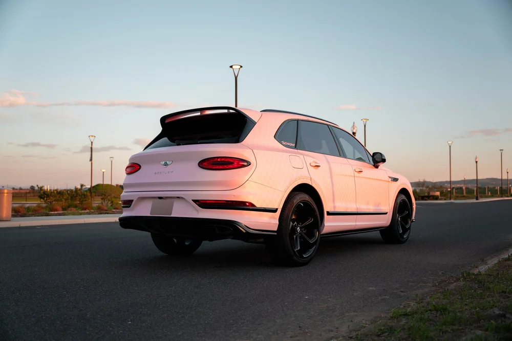 Complete opposite rear three-quarter view of the pink Bentayga, showing its black wheels, roof spoiler and side body at sunset