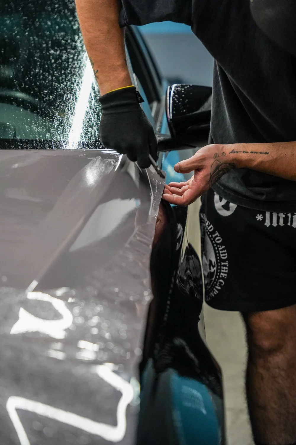 Gloved hand and tool working at a loose light-grey vinyl edge beside wet glass and a side mirror