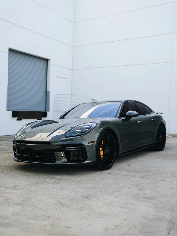 Gloss dark Porsche Panamera in a complete front three-quarter view with black wheels and yellow brake calipers outside a white building