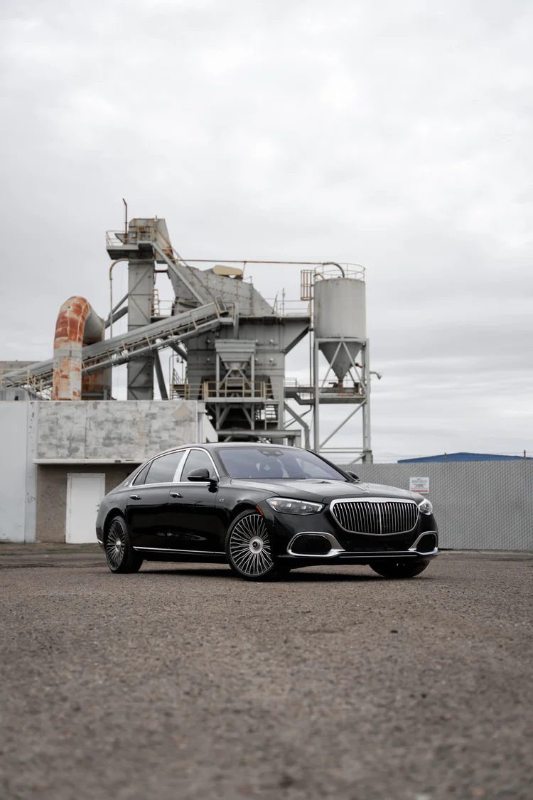 Tall complete front-three-quarter portrait of the black Maybach centered low beneath a cloudy sky and industrial towers