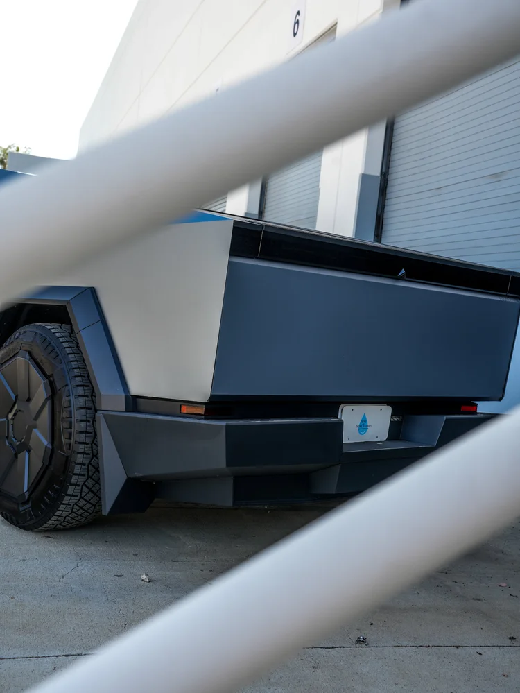 Tesla Cybertruck tailgate, Toyz plate, rear bumper and rear-wheel detail viewed through two foreground rails.