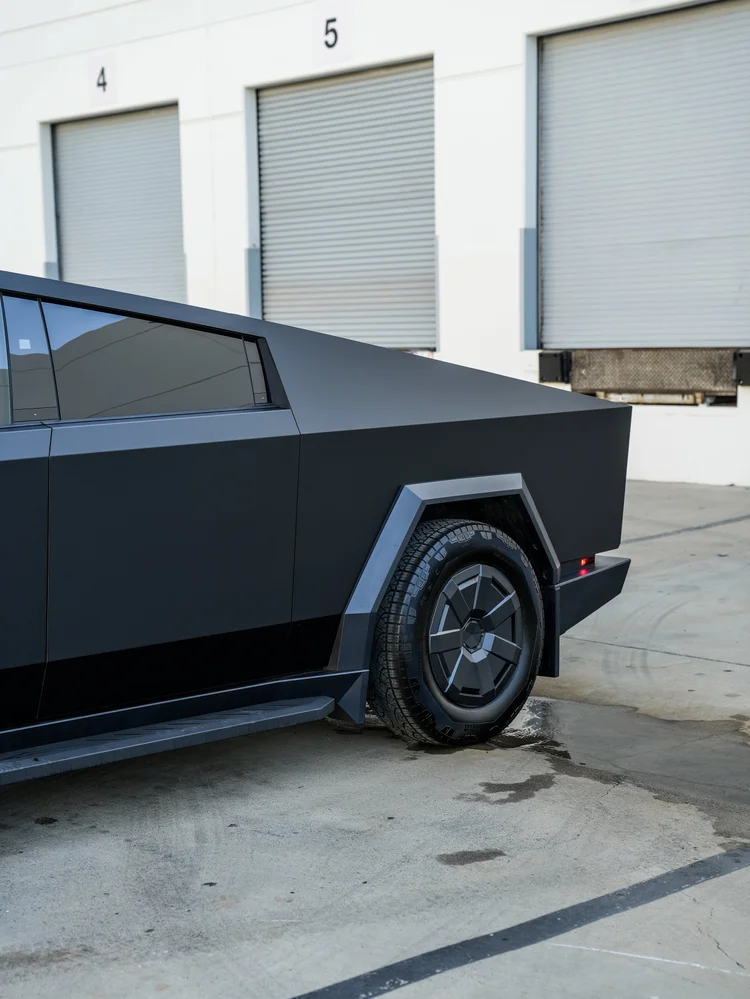 Tesla Cybertruck rear-quarter detail showing matte black side panels, dark side glass, the rear wheel and angular wheel-arch trim.