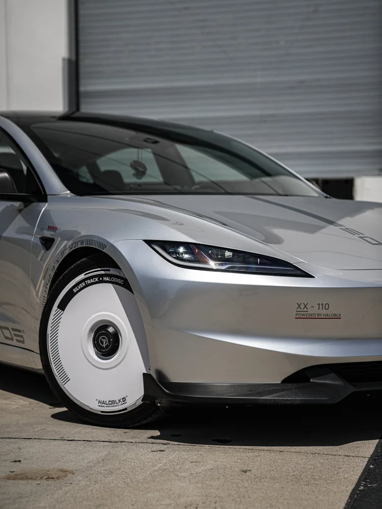 Close view of the silver Model 3 front headlamp, fender, bumper and white disc-style wheel cover beside a grey roller door.