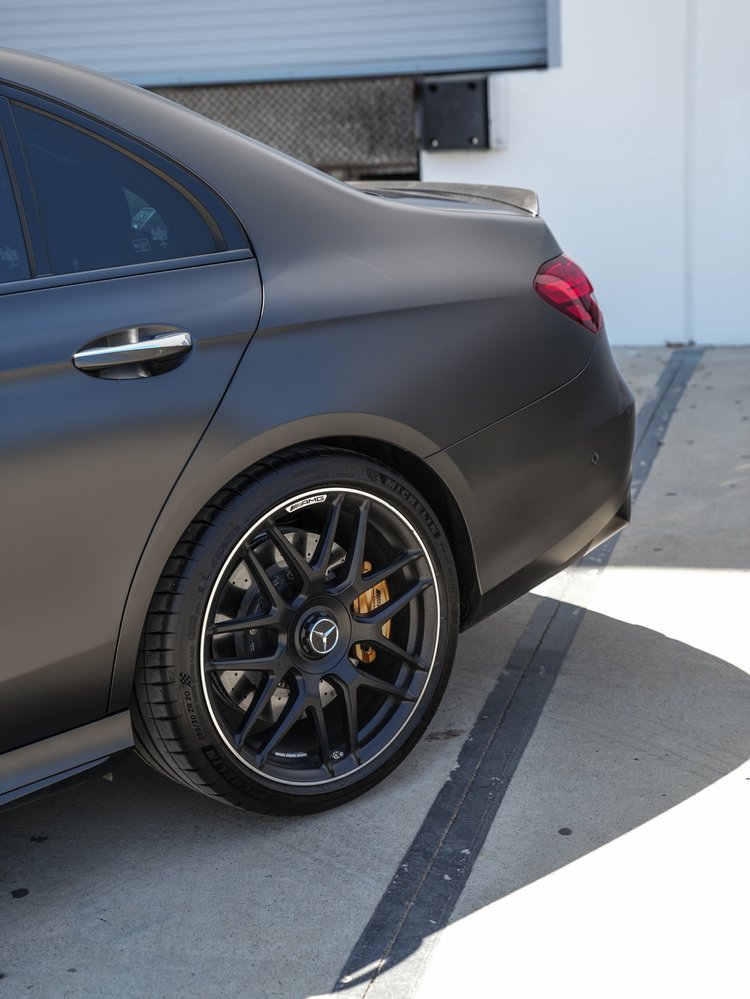 Mercedes-AMG E 63 S rear-wheel and rear-fender detail showing dark spokes, a yellow-gold caliper, red taillamp, rear-door handle and rear lip.