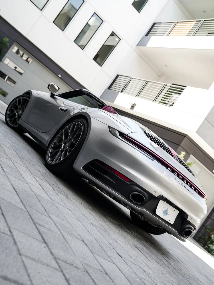 Tilted low rear three-quarter view of the silver open-roof Porsche 911 with dark multi-spoke wheels, twin round exhaust outlets and a red rear lamp strip on courtyard paving.