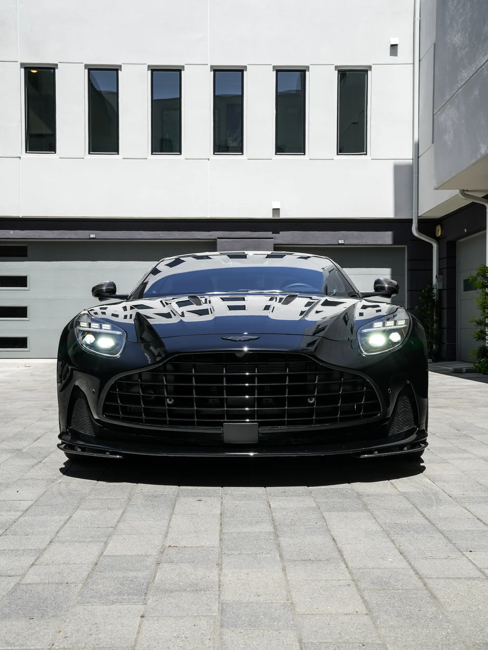 Straight-on front view of the black Aston Martin DB12 with illuminated headlamps and a wide grille.