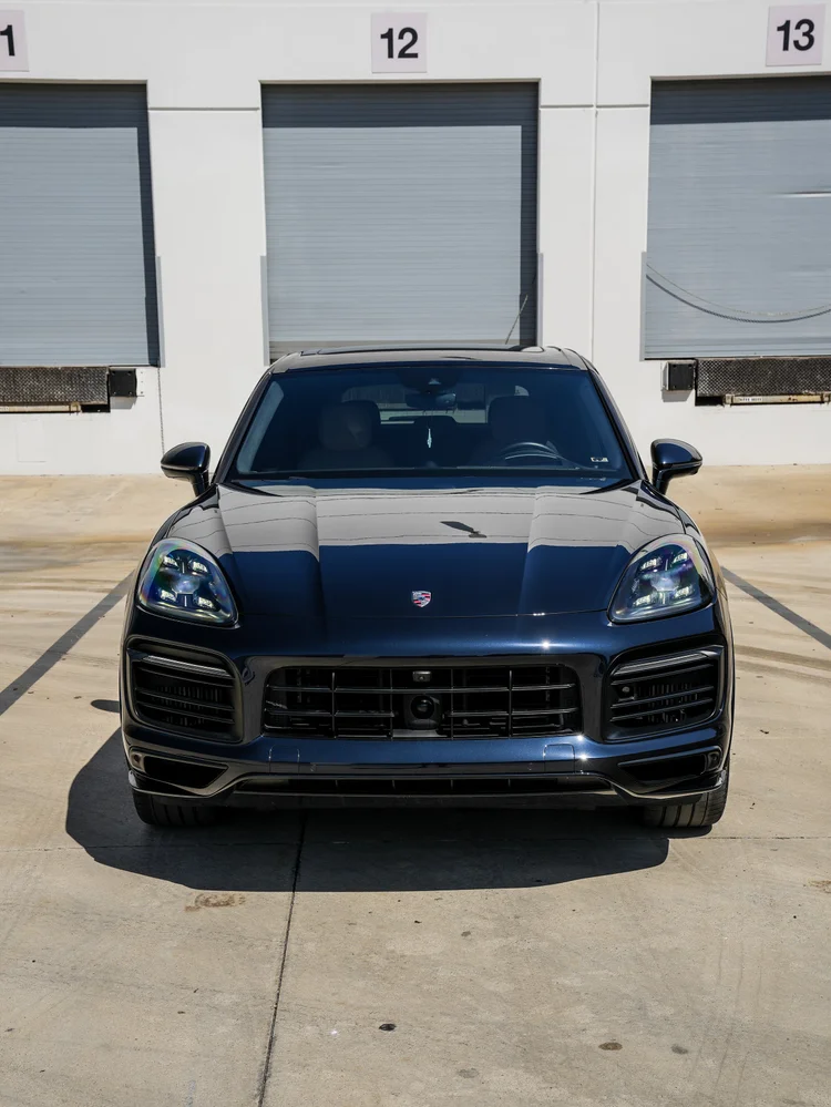 Complete straight-front view of the Porsche Cayenne GTS with its hood badge, black grille and headlamps outside workshop doors twelve and thirteen.