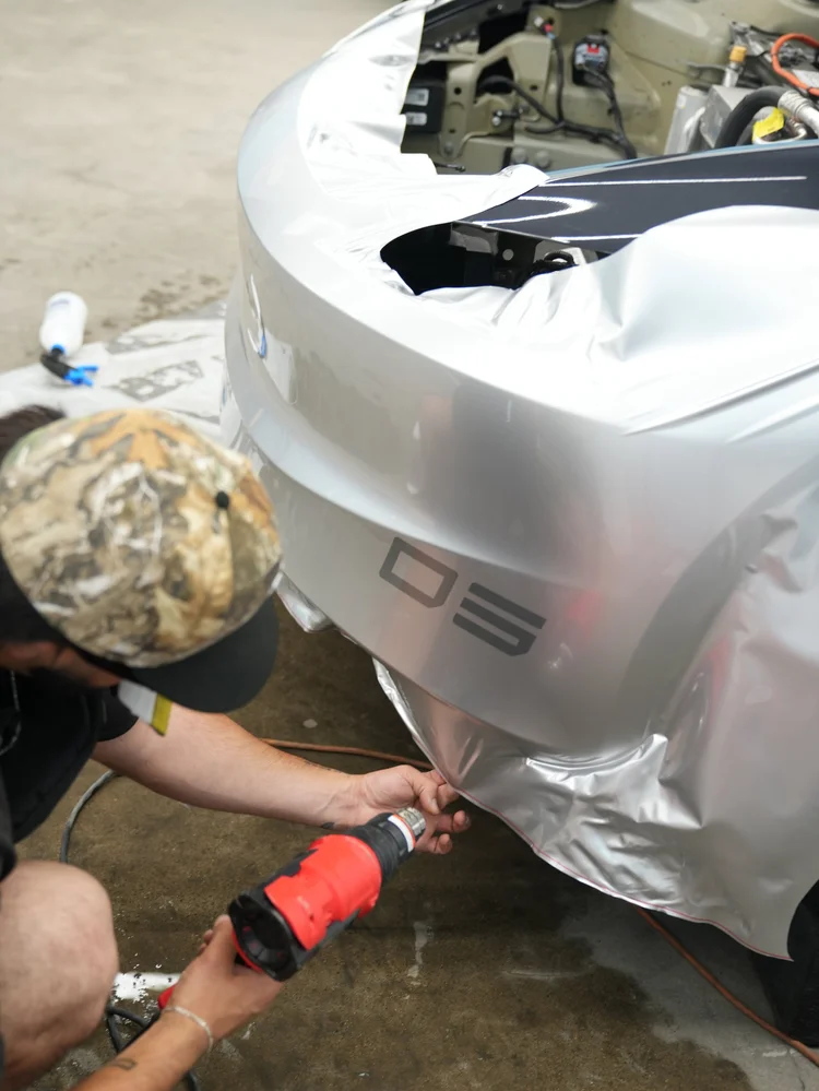 Installer holding silver film at the Model 3 lower front bumper while using a red heat gun, with loose film around the open front area.