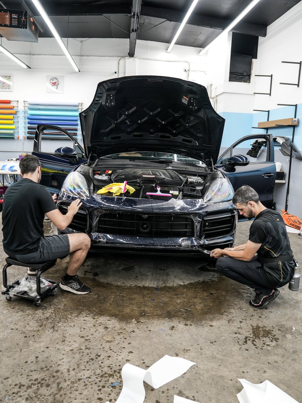 Two technicians handling wet clear film around the Porsche Cayenne GTS front bumper with its hood and front doors open inside the workshop.