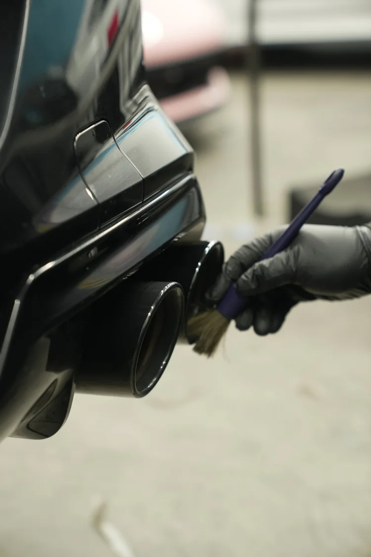 Gloved hand using a small brush around one of the BMW M8 rear exhaust outlets beside the dark rear bumper.