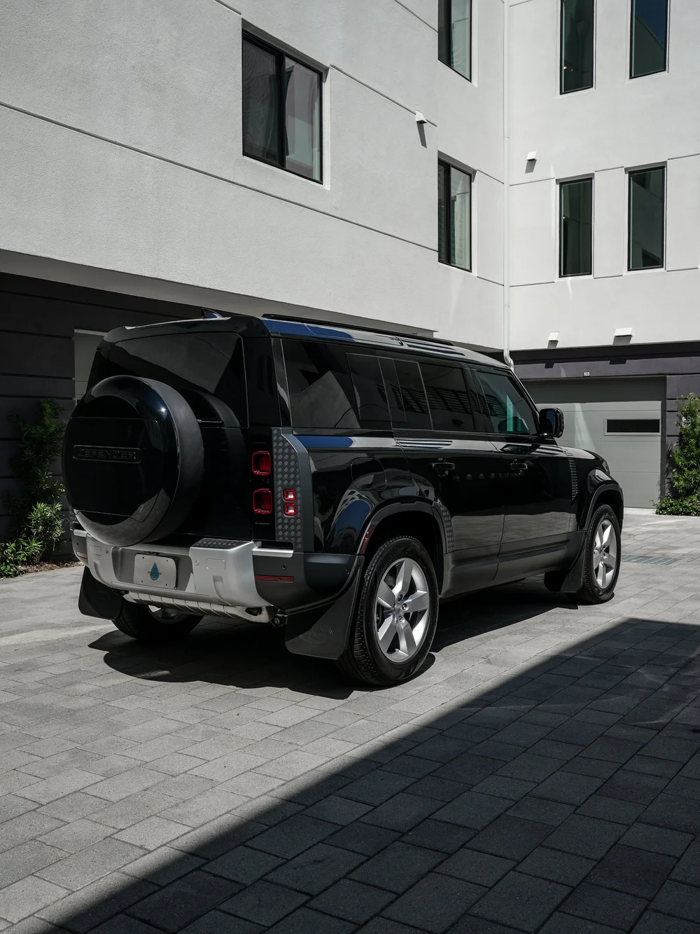 Complete rear-three-quarter view of the dark Defender with a round rear spare-wheel cover, stacked red lamps and silver wheels outside an industrial building
