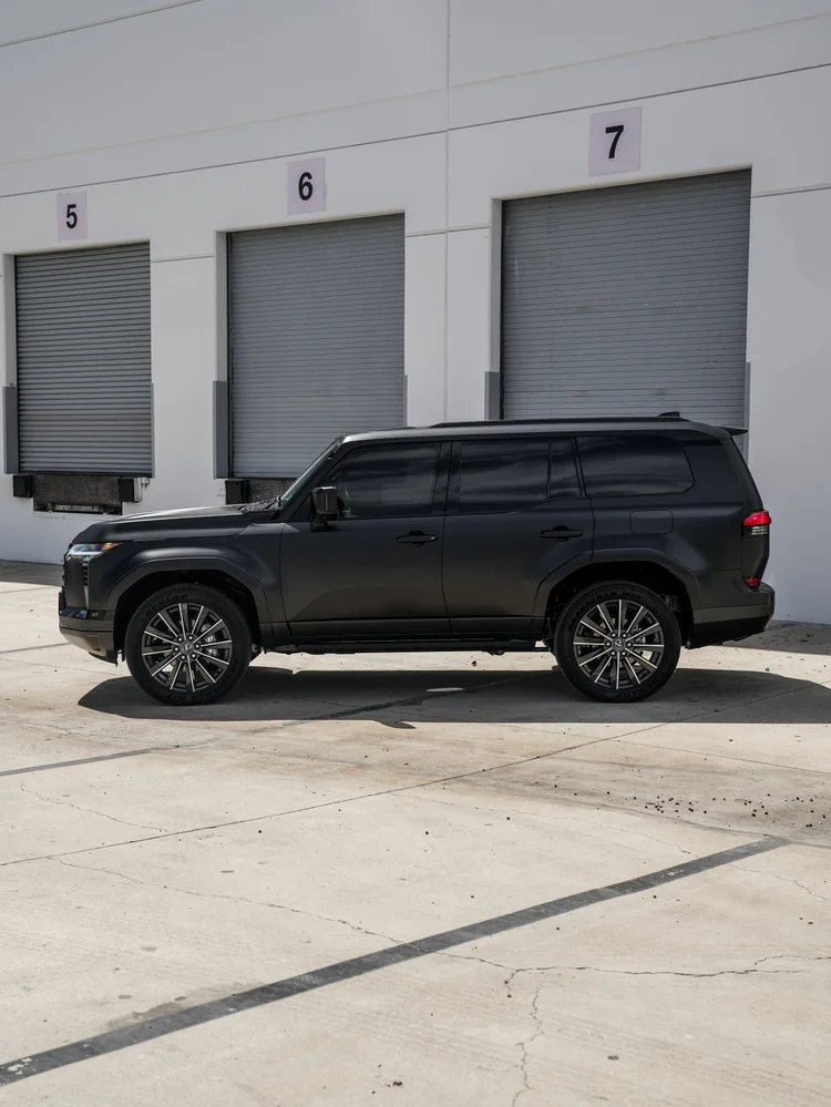 Complete side profile of the dark GX 550 with bright multispoke wheel faces, roof rails, dark side glass and a long running board beside garage doors