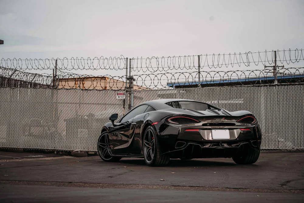 Whole McLaren 570S viewed from the rear corner beside a tall chain-link fence.
