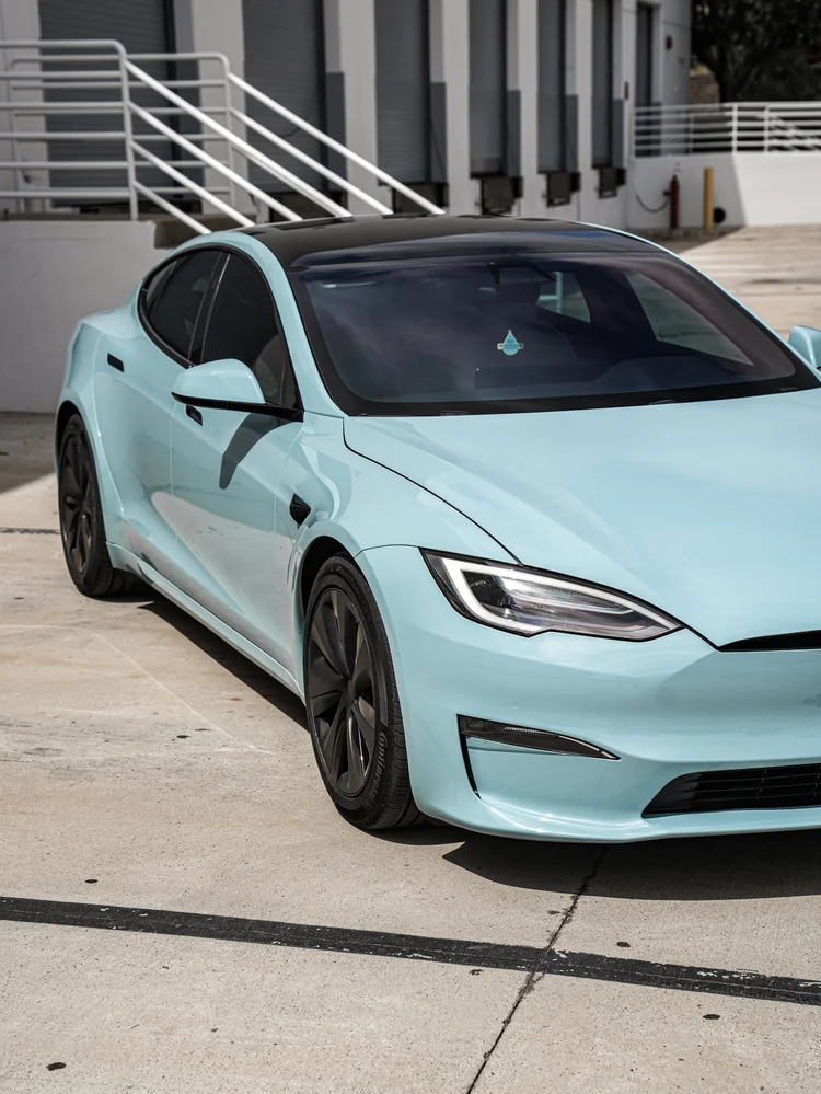 Pale blue Tesla Model S in a partial front three-quarter view beside white stairs, with the nose cut by the right image edge.