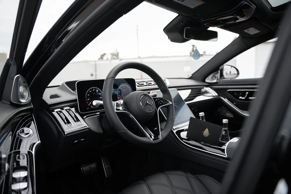 Dark Maybach cabin through the open driver door with a Mercedes-star steering wheel, dashboard screens, bright vents and bottles on the console