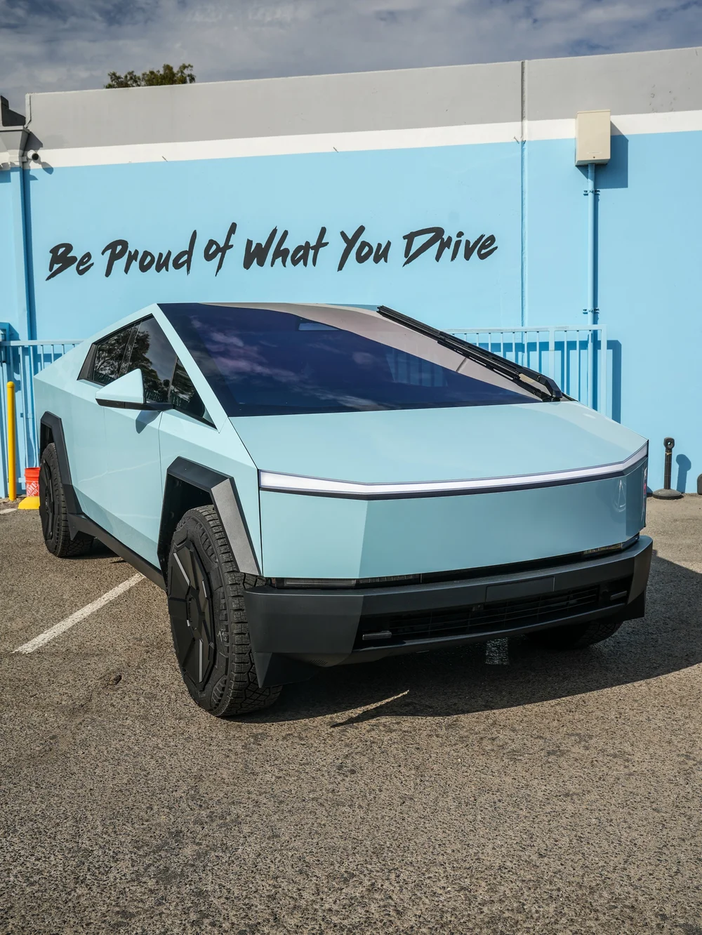 Complete front three-quarter view of the pale blue Cybertruck in front of the blue Be Proud of What You Drive wall.