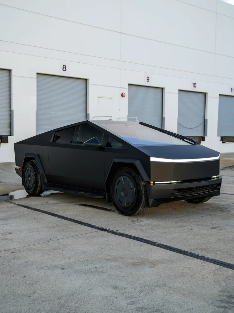 Wider right-facing front three-quarter view of the complete matte black Tesla Cybertruck beside numbered workshop doors.