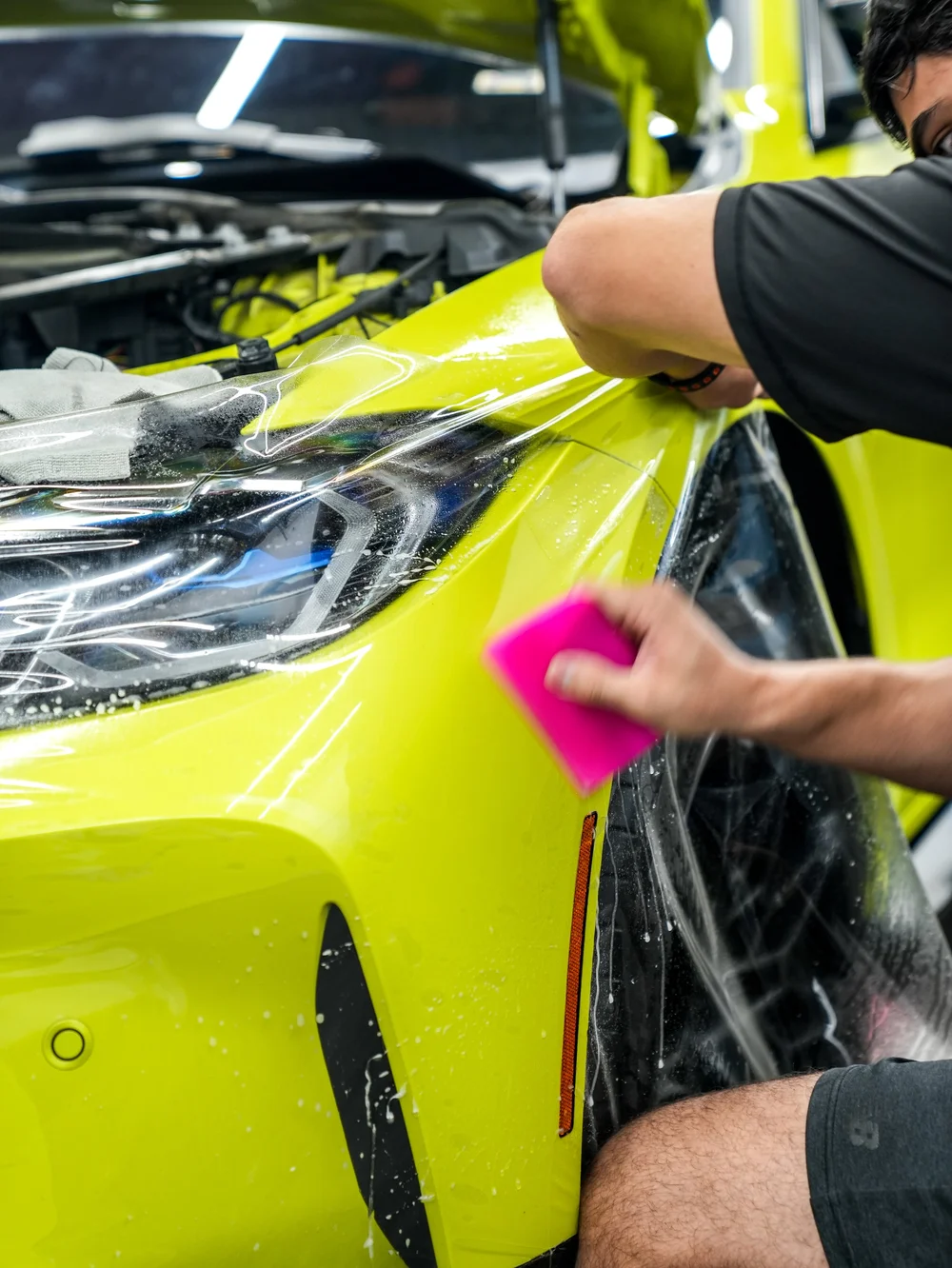 Technician handling wet transparent film at the BMW M4 headlight and front corner, with a pink flat tool against the yellow-green panel beside the front wheel.