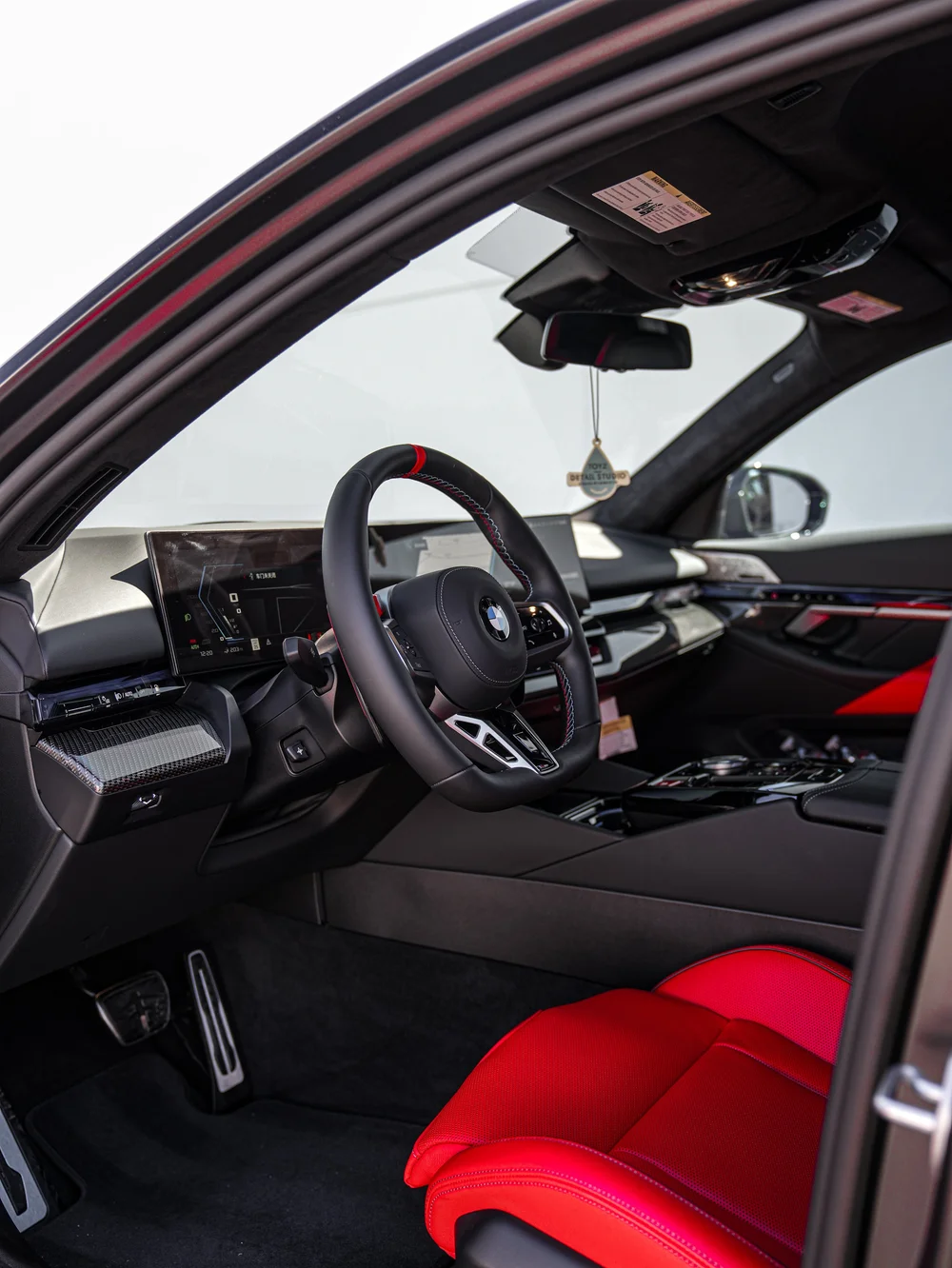 BMW G90 M5 cabin viewed through the open front door, showing the steering wheel, dashboard and red seat
