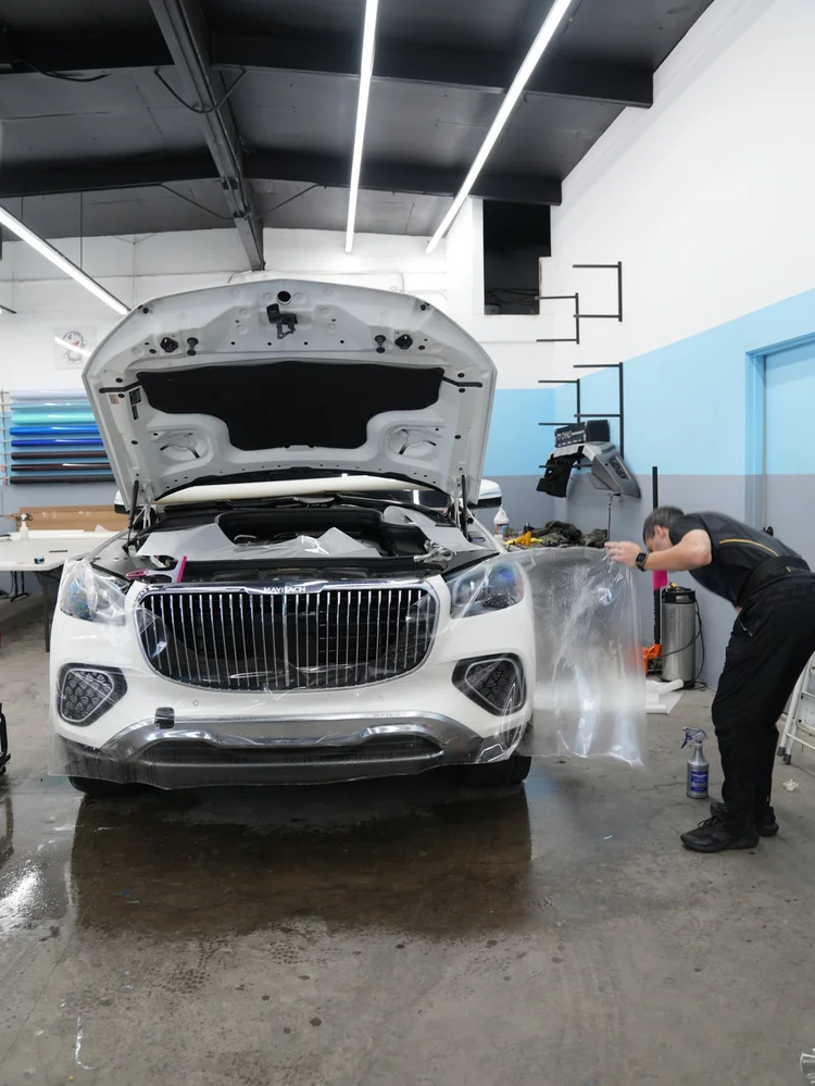 Wide frontal workshop view with the hood open and a technician positioning wet clear film across the white front bumper and headlamp areas.