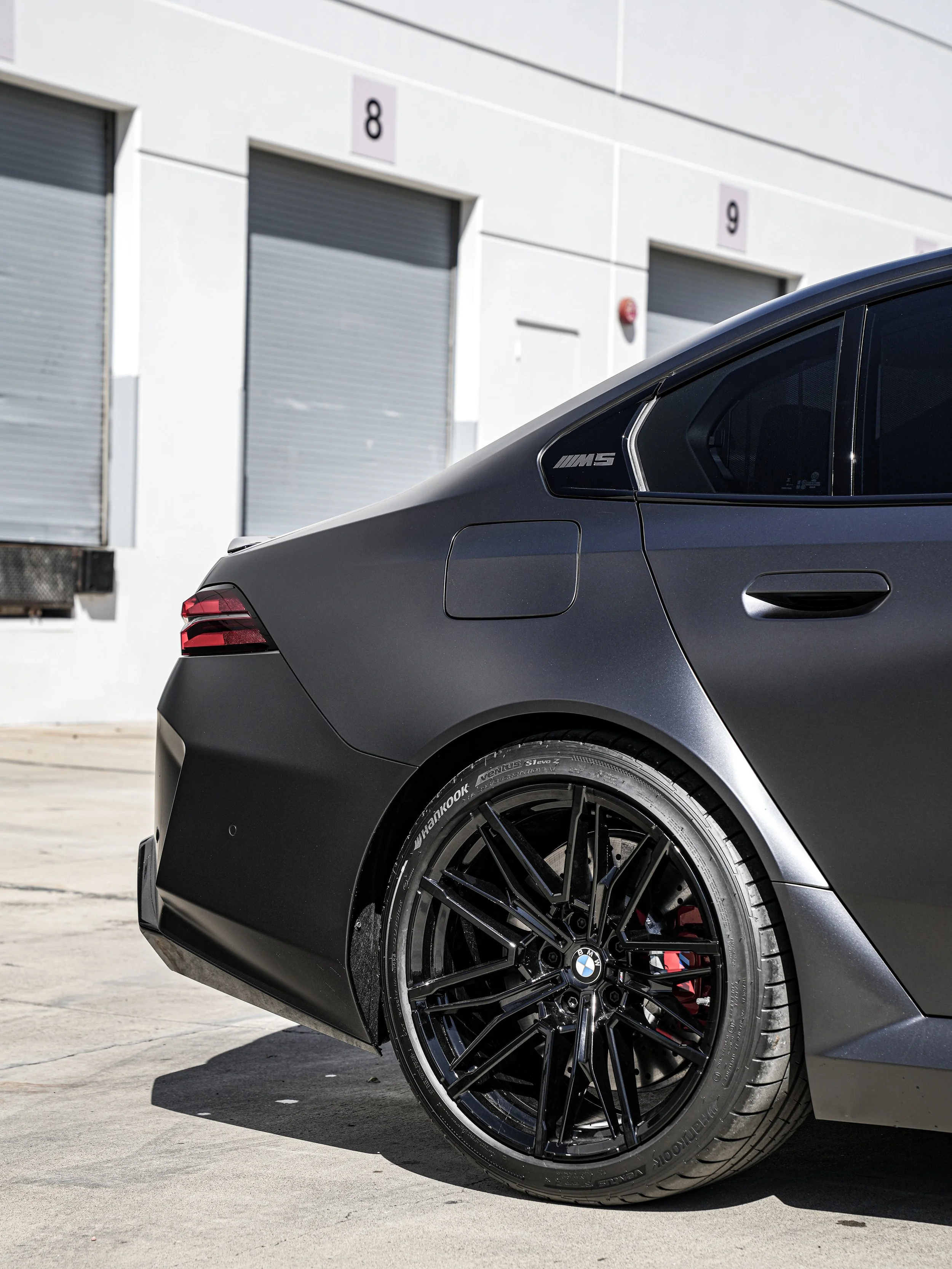 Close-up of the rear side of a matte black BMW M5 with black alloy wheels, red brake calipers, and an 'M5' badge, parked outside a building with numbered garage doors.