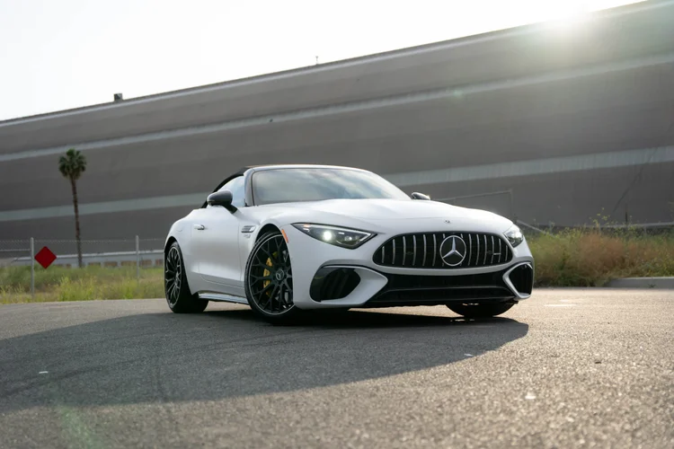 Complete low front-three-quarter view of a white Mercedes-AMG SL 63 with illuminated headlamps, dark wheels, yellow calipers and a dark fabric roof on pavement.