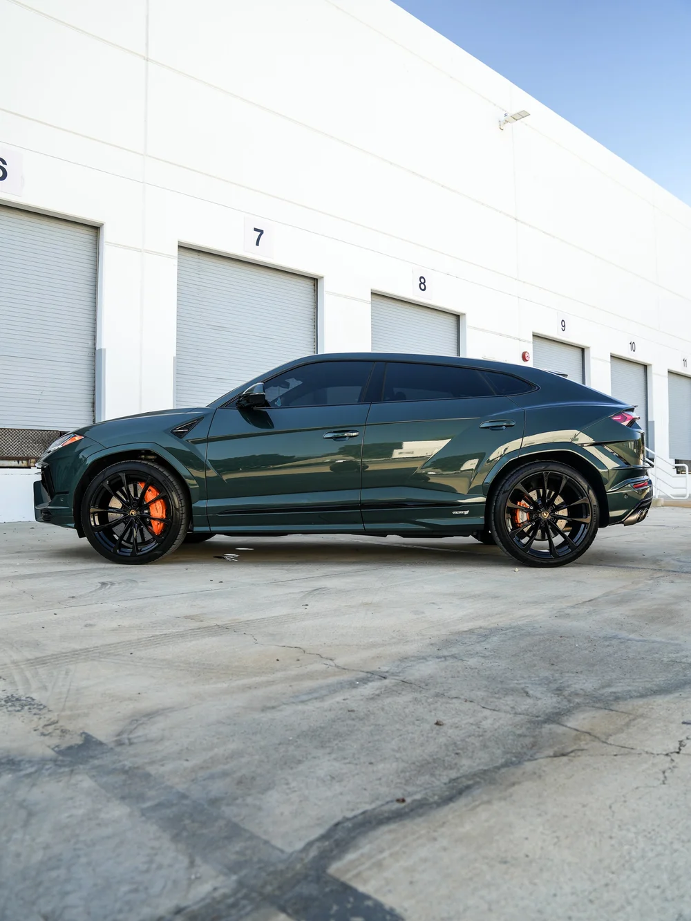 Complete closed-door side profile of the dark green Lamborghini Urus S facing left, with black multi-spoke wheels, orange brake calipers and dark side glass beside a white building.