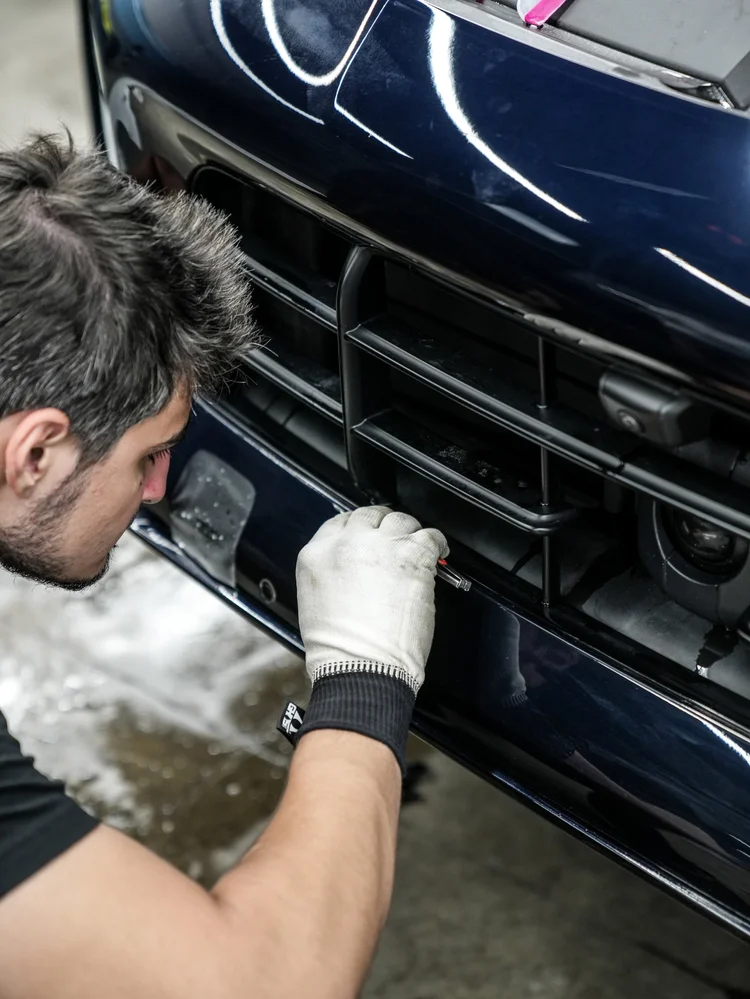 Gloved hand holding a slender tool near the Porsche Cayenne GTS lower front grille and bumper edge.