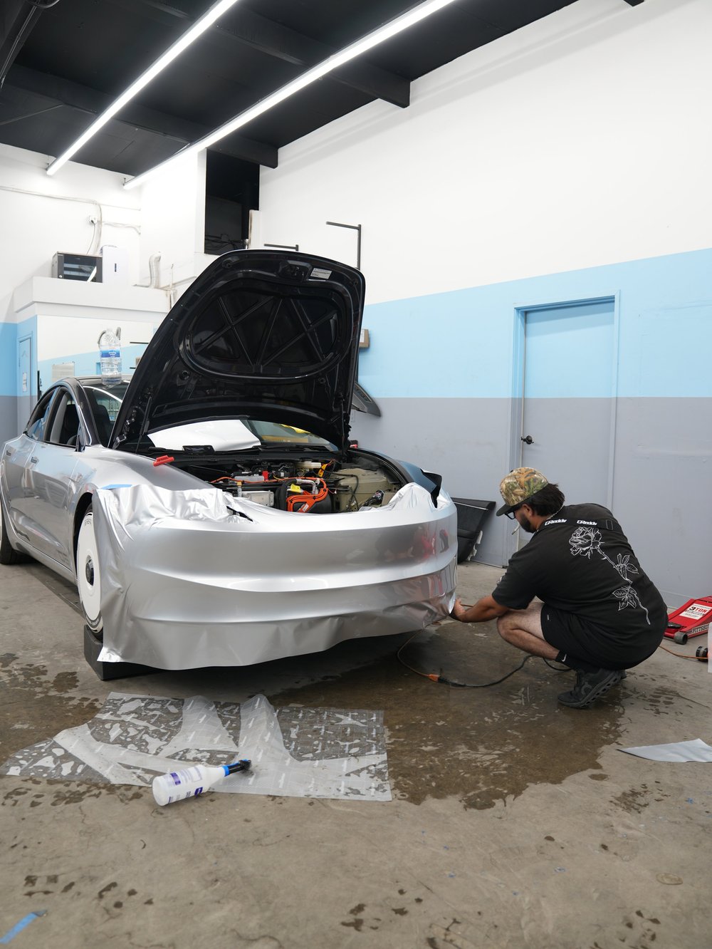 Installer kneeling beside loose silver film across the Model 3 front bumper, with the front compartment open and film scraps on the workshop floor.