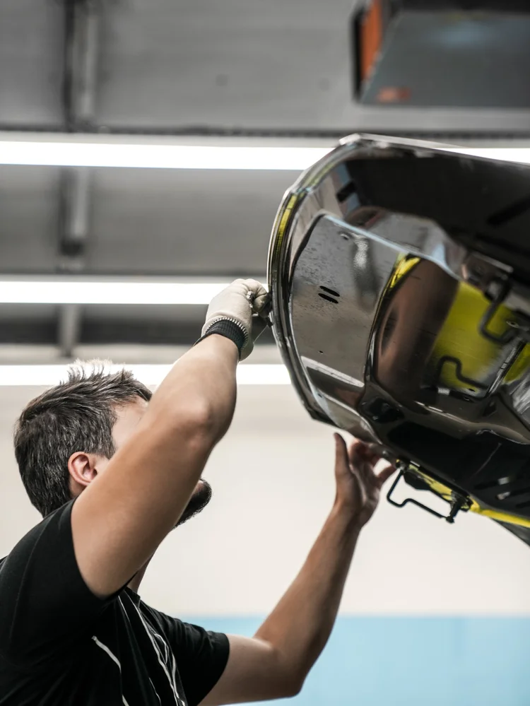 Technician reaching with gloved hands and a small tool beneath the lip of the Porsche Cayenne GTS raised hood.