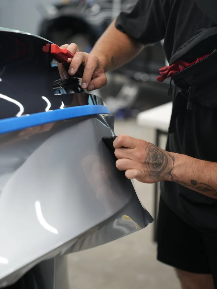 Installer hands smoothing silver film around a dark curved body-panel edge beside blue tape with a small hand tool.