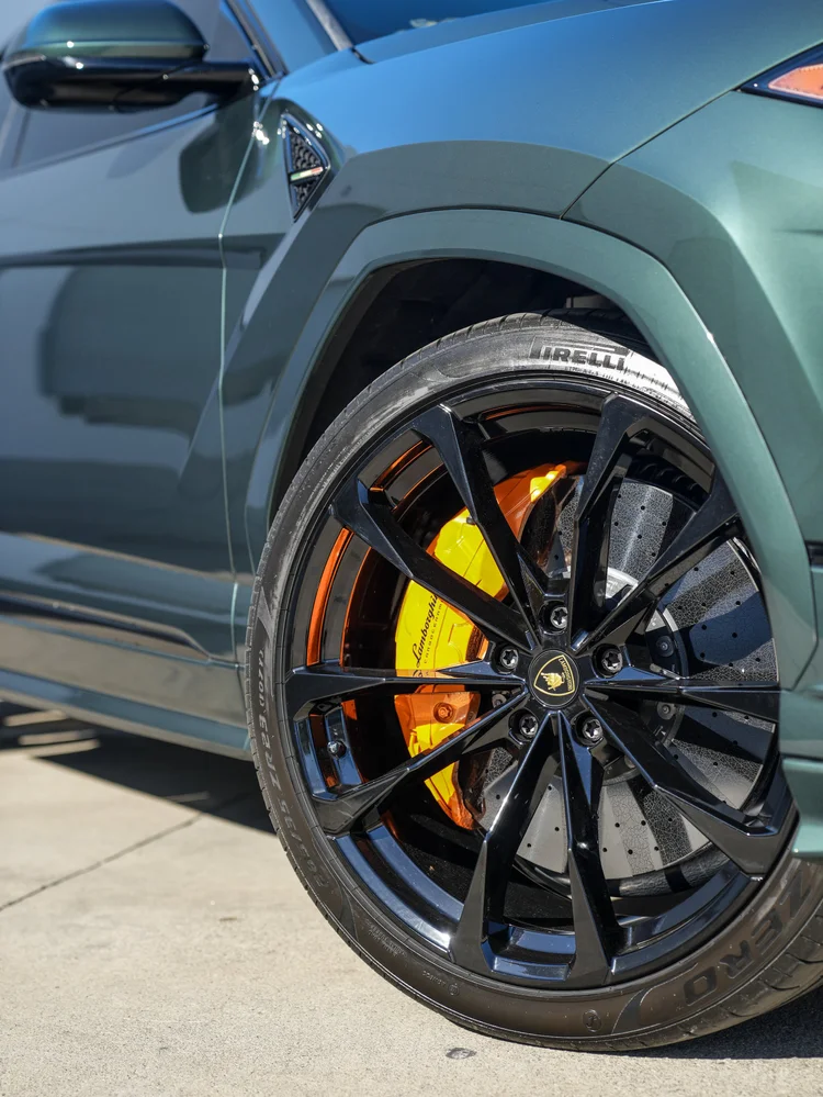 Black multi-spoke front wheel of the green Lamborghini Urus S with a gold bull center emblem, visible Pirelli tire lettering and an orange Lamborghini brake caliper.