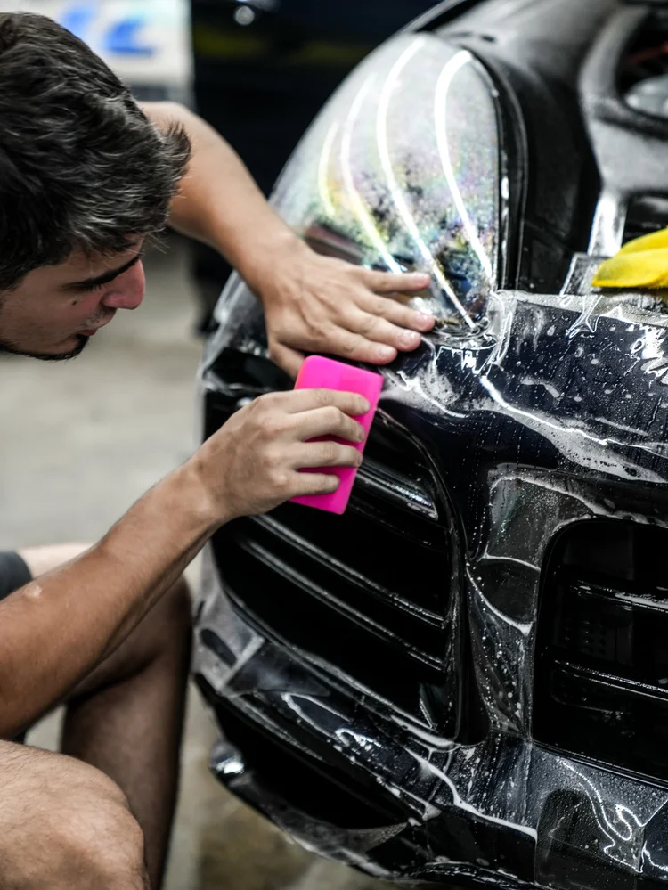 Technician holding a pink squeegee against wet clear film on the Porsche Cayenne GTS bumper below a headlamp.