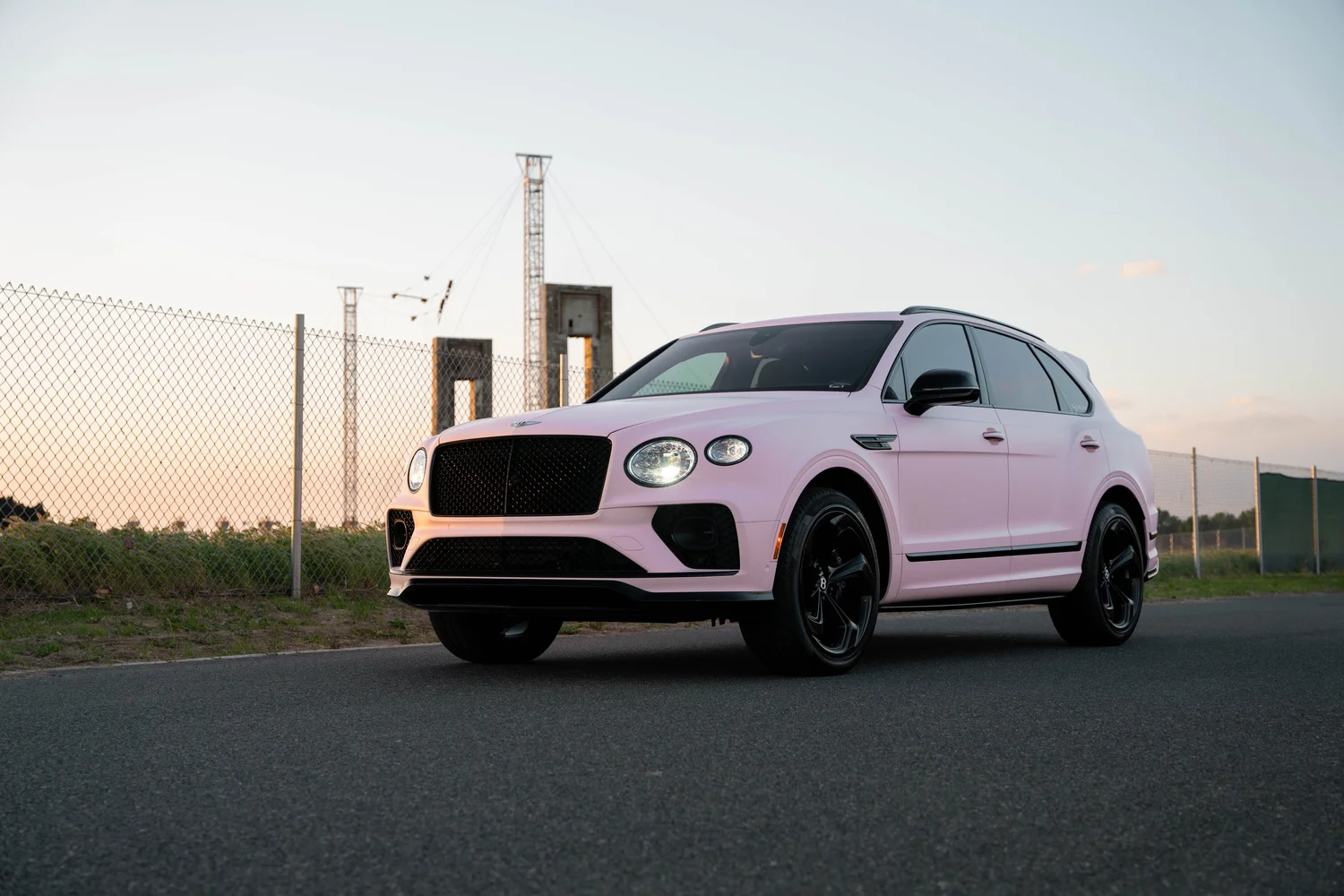 Complete front three-quarter landscape of the Sakura Pink Bentayga parked beside a chain-link fence