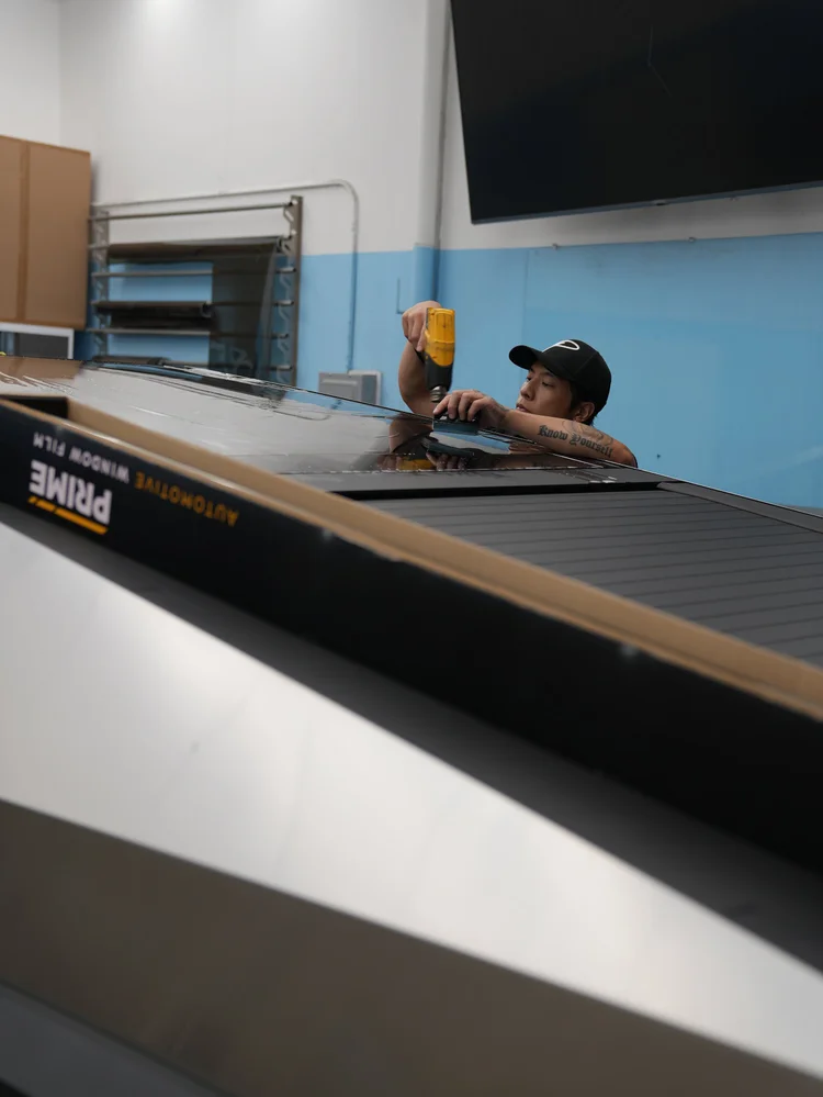 A technician holding a yellow heat tool near wet glass at roof height on the Tesla Cybertruck, with a film box visible nearby.