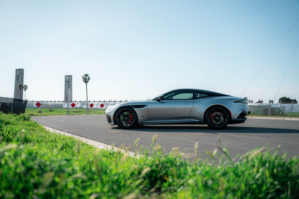 Matte-finish Aston Martin DBS in a full side profile on the road beside low green plants