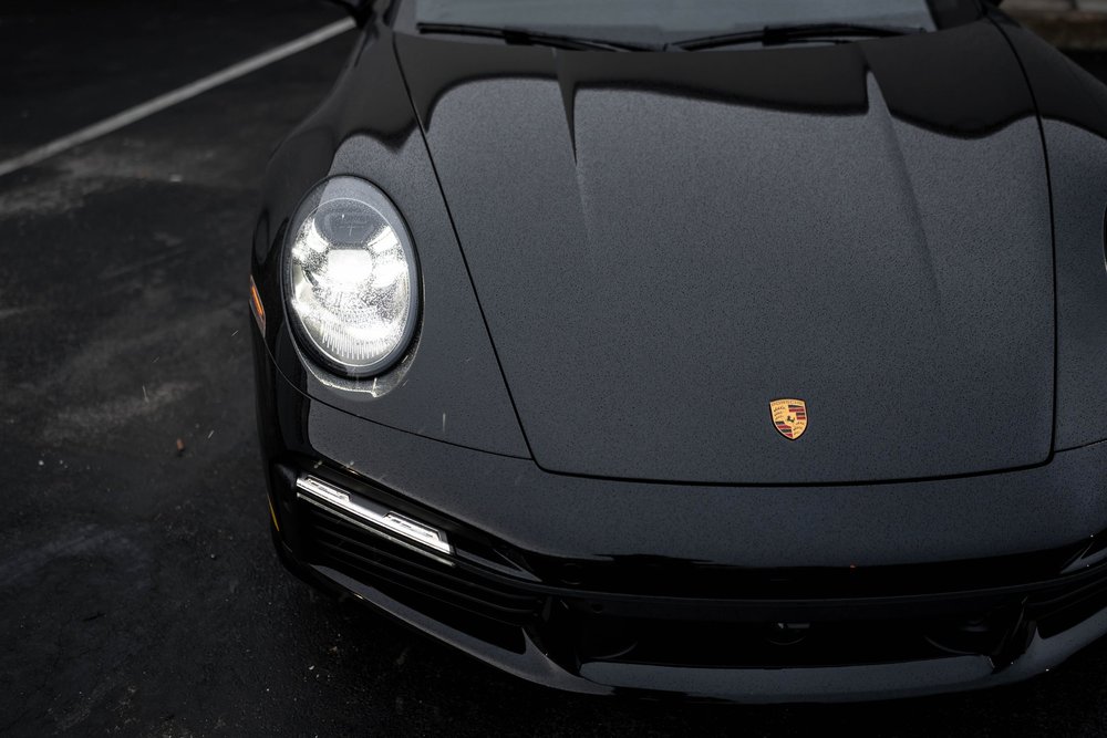 High front detail of the black Porsche 911 Turbo S hood and illuminated headlight with water droplets