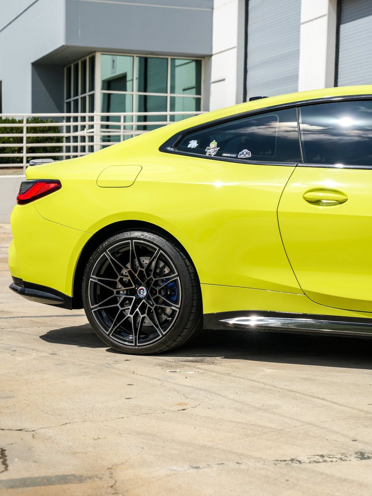 BMW M4 rear wheel and blue brake caliper beneath the yellow-green rear quarter and door, with dark side glass and part of a red rear lamp beside the shop entry.