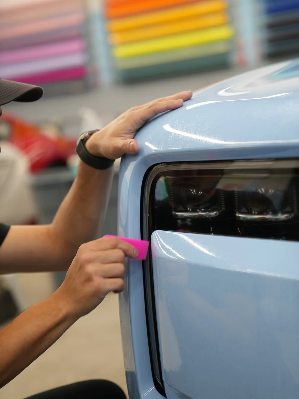 Technician using a pink squeegee on clear film beside the Cullinan headlight and Powder Blue front bodywork