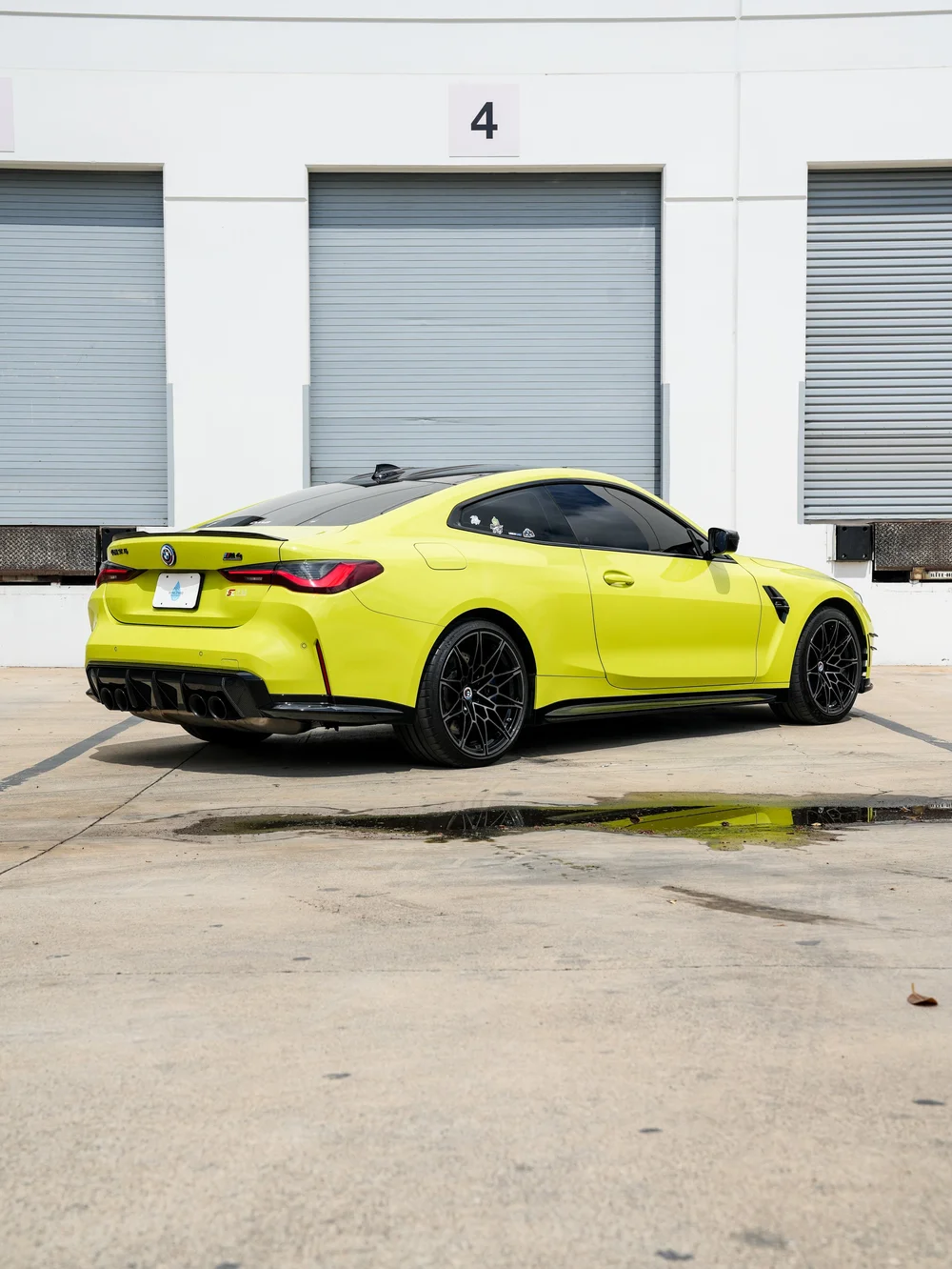 Complete rear three-quarter view of the yellow-green BMW M4 facing right, with a dark roof, black wheels, rear spoiler, red taillights and closed doors outside gray workshop doors.