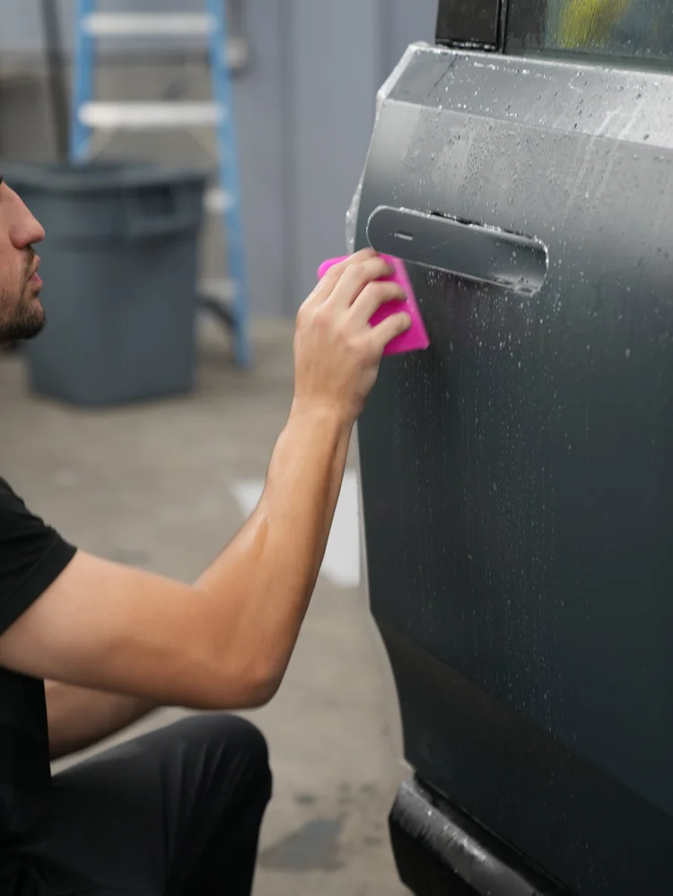 A technician pressing a pink squeegee against wet film on a Rivian R1T door panel below its flush handle.