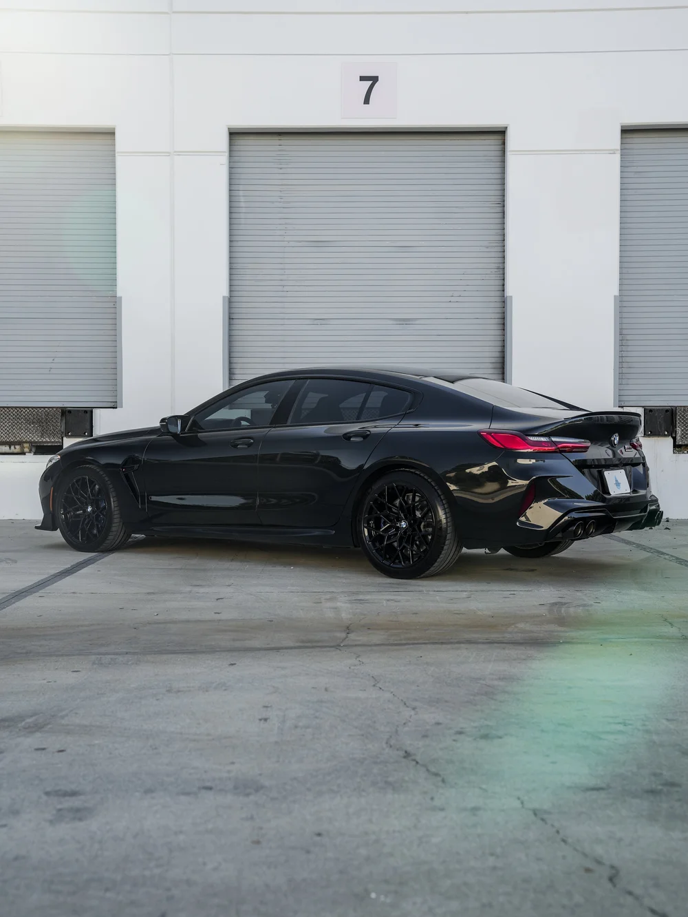 Complete left-facing rear three-quarter view of the dark BMW M8 with red taillamps, dark wheels and four exhaust outlets outside workshop door seven.
