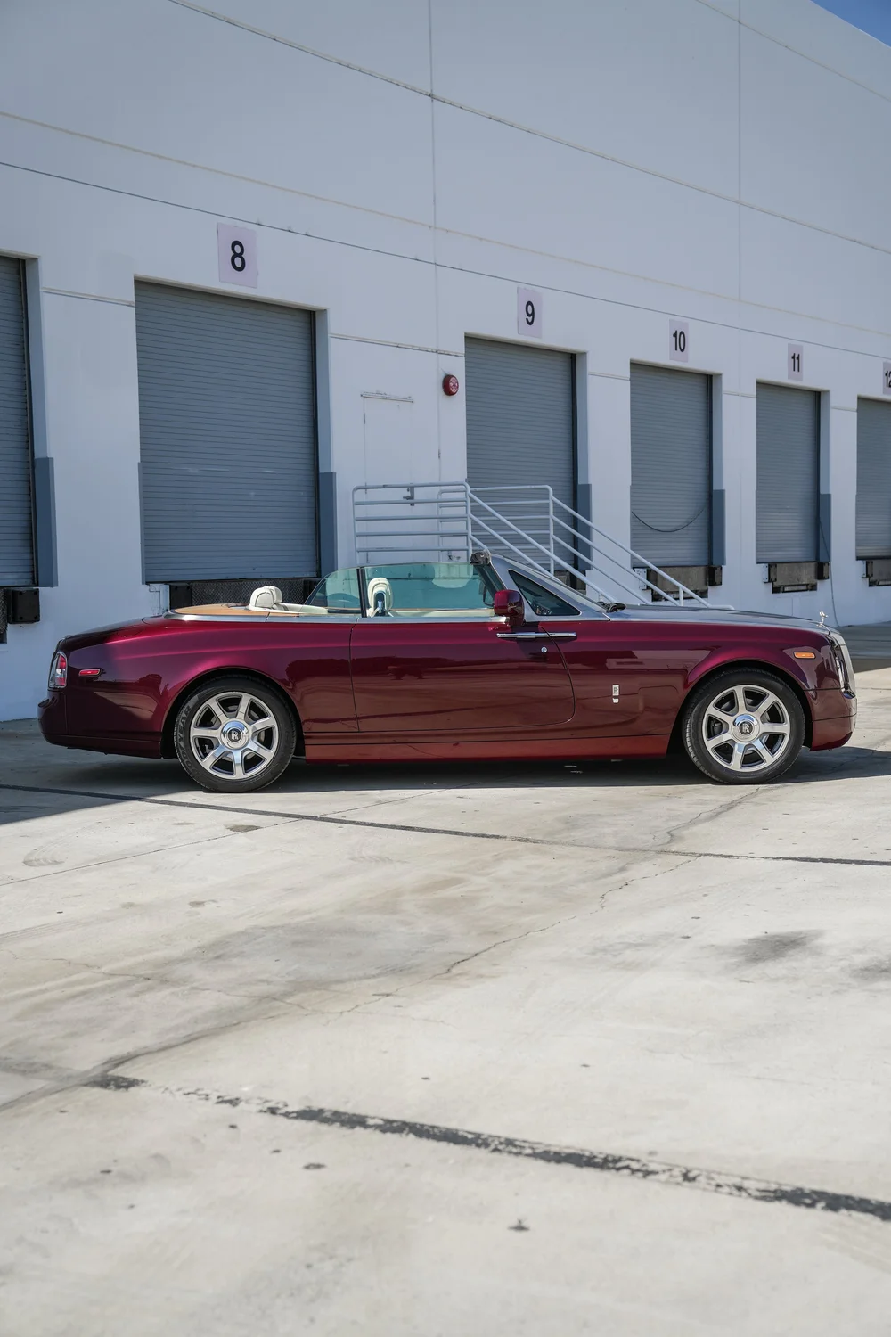 Complete right-facing side profile of the burgundy Rolls-Royce Phantom Drophead Coupé with its roof lowered, cream-colored headrests, silver wheels and contrasting silver-colored hood outside numbered workshop doors.