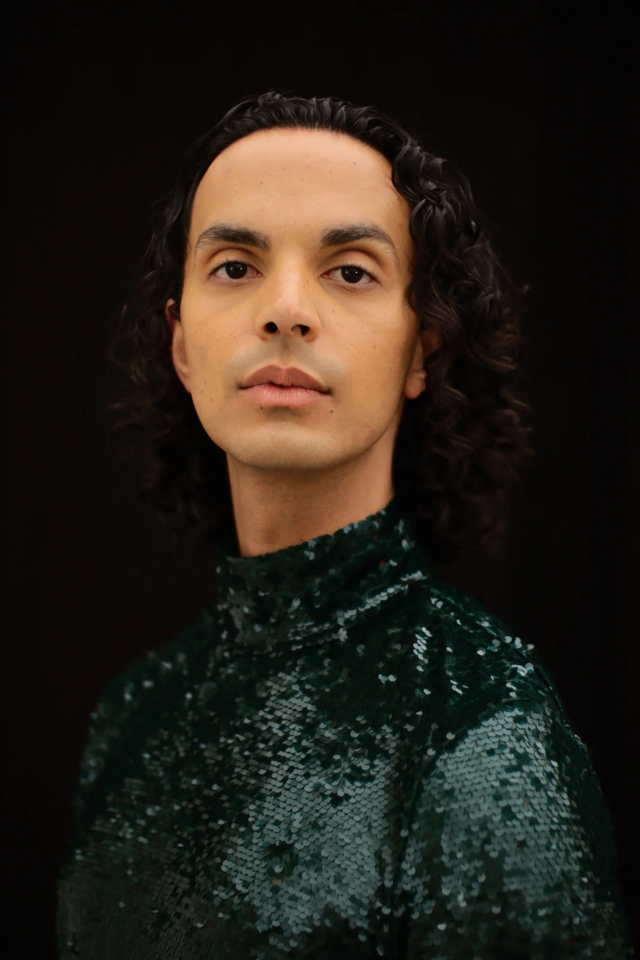 Portrait of a person with curly dark hair wearing a dark green sequin turtleneck against a black background.