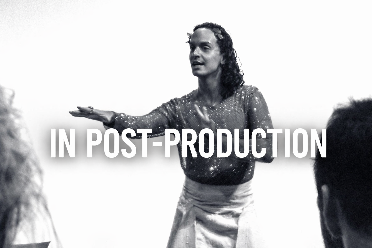 A woman in a glittery top and skirt is speaking passionately, with her arms outstretched, during a presentation or discussion, with the words 'IN POST-PRODUCTION' overlaid.