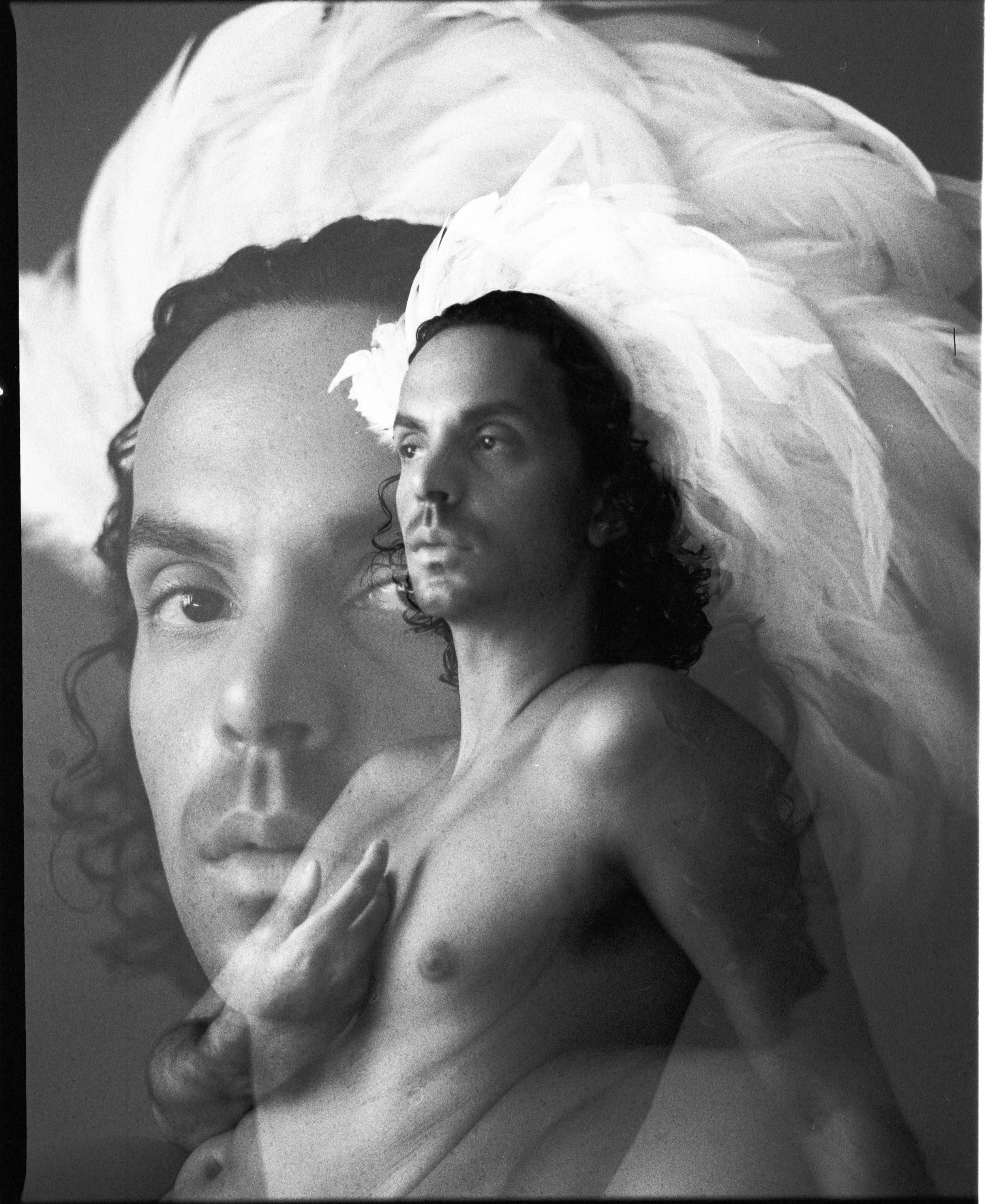 Black and white photograph of Marcelo Kuna with a large feathered headdress. Marcelo has curly hair and a contemplative expression, with their hand resting on their lap. The image has artistic, vintage quality.