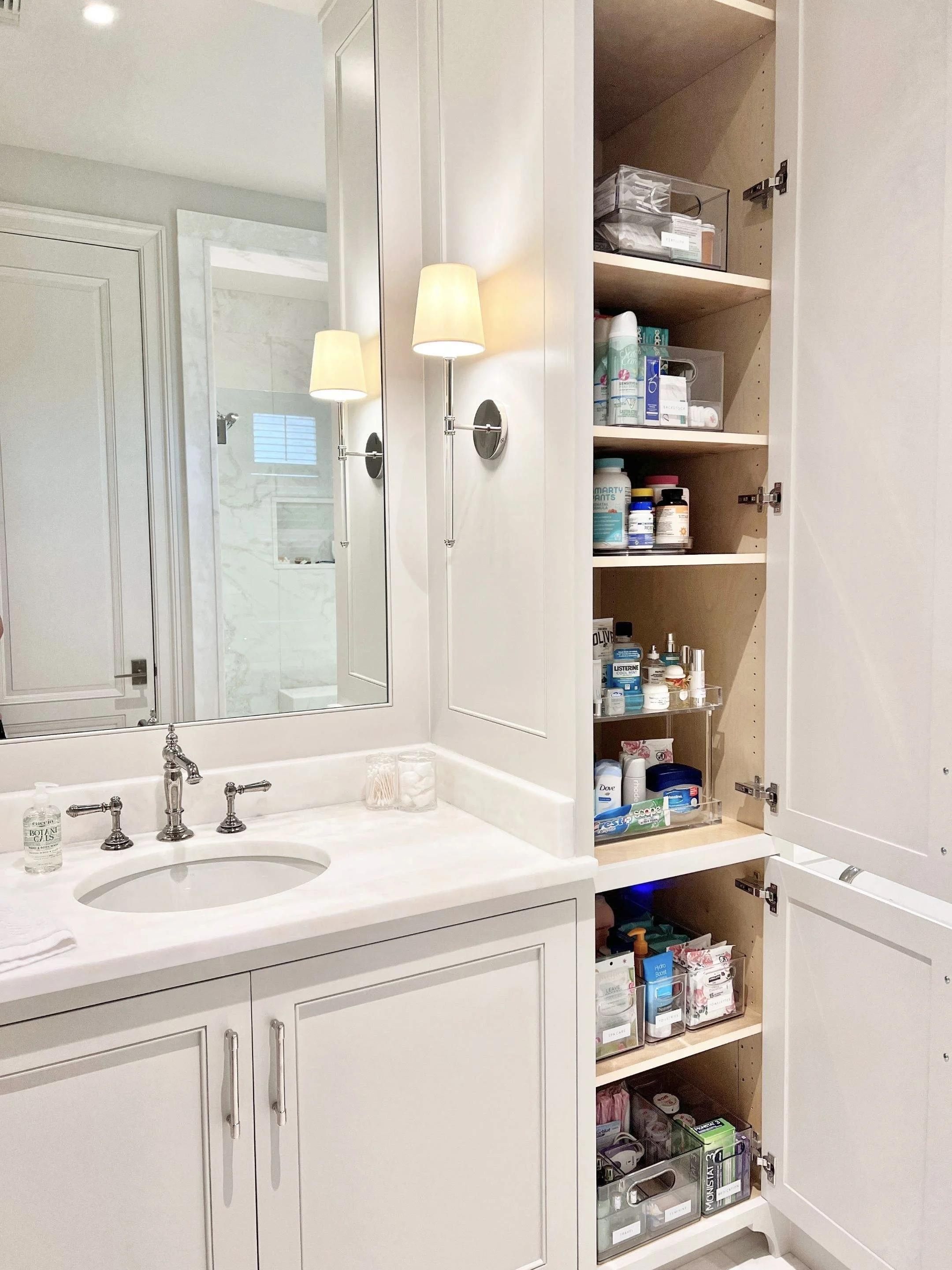 Bathroom with a white vanity, marble countertop, and a mirror. Open bathroom cabinet filled with toiletries and supplies, with a wall-mounted light fixture.